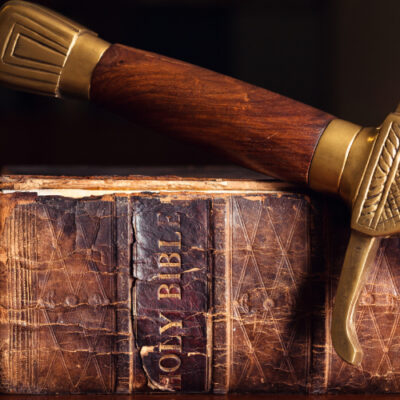 An old tattered bible sits on a table with a sword draped across the spine