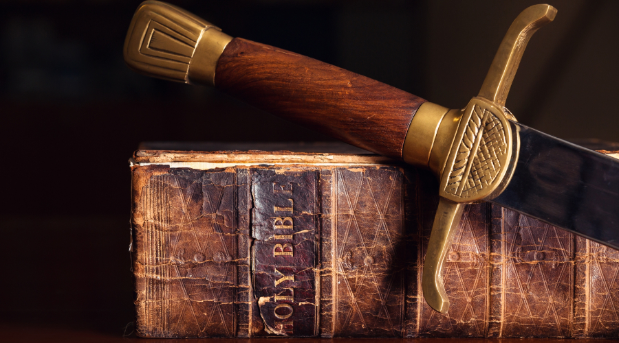 An old tattered bible sits on a table with a sword draped across the spine